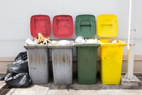A row of four large, wheeled rubbish bins with closed colored lids in red, red, green, and yellow, situated outdoors on a paved surface against a white wall. The grey bin on the left is filled with waste and has a black rubbish bag leaning against it. The waste in the bins appears to include plastic packaging, paper, and mixed refuse, creating a cluttered appearance. The green bin in the middle is also filled with waste, partly spilling over the top, and the yellow bin on the right is closed. The scene suggests a waste collection or disposal area, typical for commercial or residential waste management in Bow, with the bins positioned directly on the pavement and a small drainage grate nearby. The overall setting conveys an organized but utilitarian environment focused on rubbish storage and removal, consistent with services provided by Commercial Waste Removal Bow for local businesses or properties.