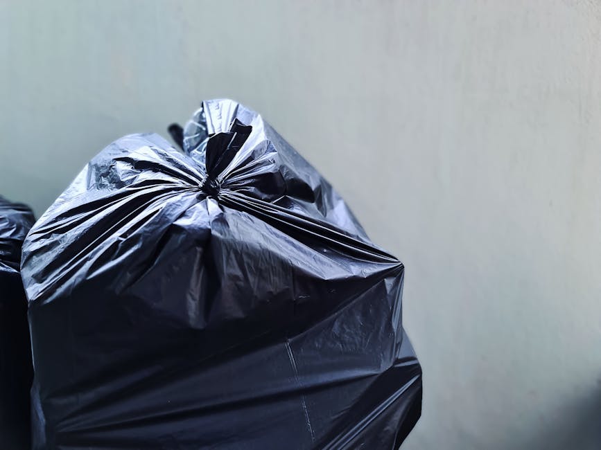 A black wheelie bin with the label 'ST. JOHN'S' is positioned on the pavement along a quiet street at night. The bin is filled with various waste items, including cardboard and plastic packaging, some