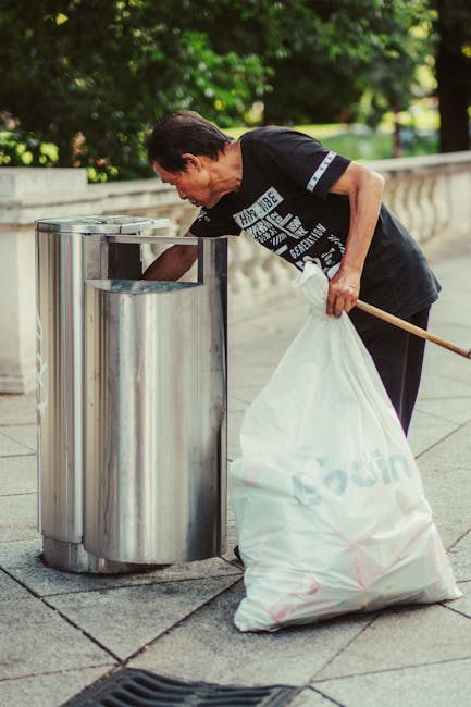 A middle-aged man with dark hair, dressed in a black T-shirt with white text and dark trousers, is seen disposing of rubbish into a tall, cylindrical metal public litter bin on a paved outdoor walkway