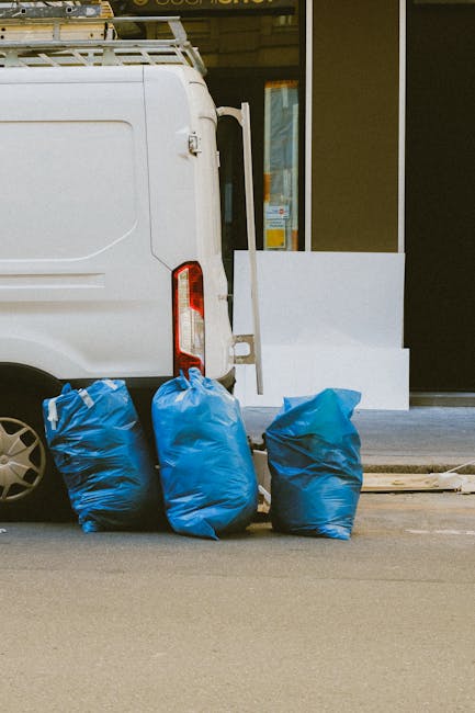 A sanitation worker in a red and yellow uniform is seen standing beside the rear opening of a large red rubbish collection truck, which is parked on the side of a road. The worker appears to be loadin