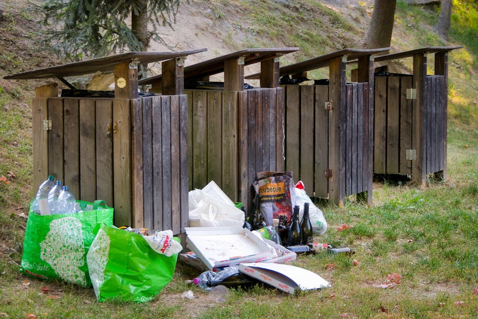 A man with dark hair, wearing a black T-shirt with white text and black trousers, is seen leaning over to dispose of a white plastic bag of rubbish into a stainless steel recycling bin on a paved side